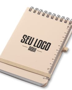Mini caderno em 3D, com capa preta e espiral, aberto e vazio. O caderno está sobre uma mesa de escritório, ao lado de um notebook, um livro e um papel em branco. A imagem transmite a ideia de organização, estudo e trabalho.